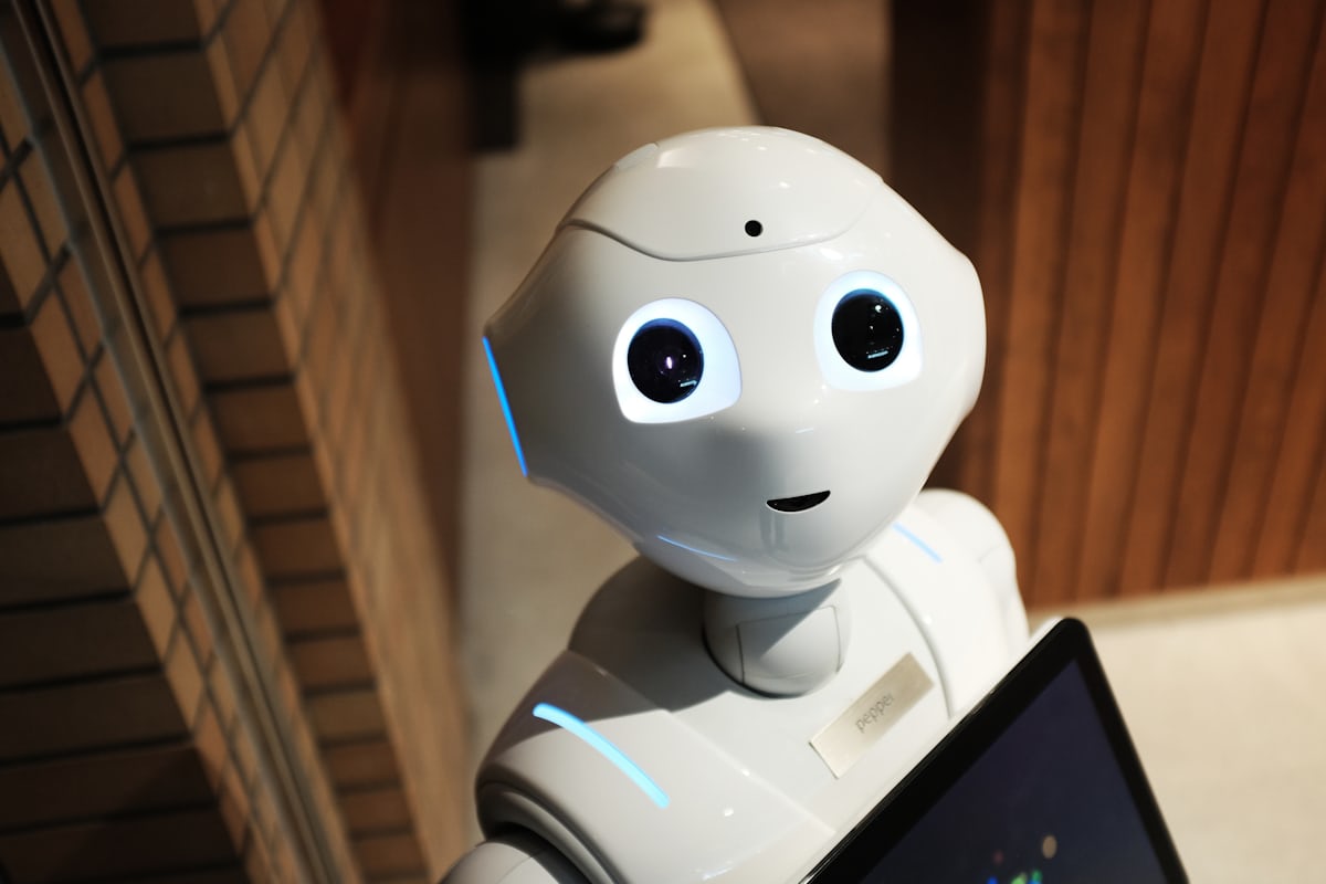 A small white humanoid robot with a screen on its chest sitting at a desk beside an open laptop.