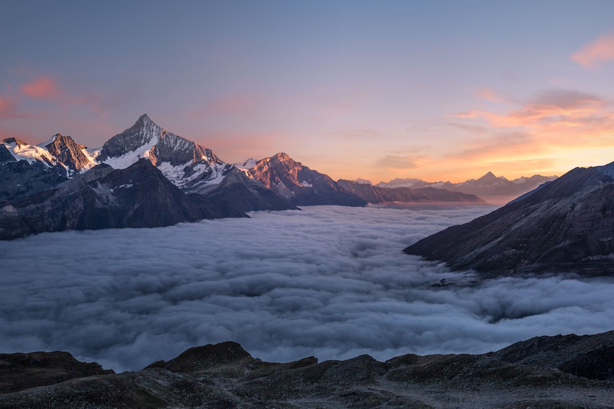 Snow-covered mountain peaks rising above a layer of cloud in low sunlight.
