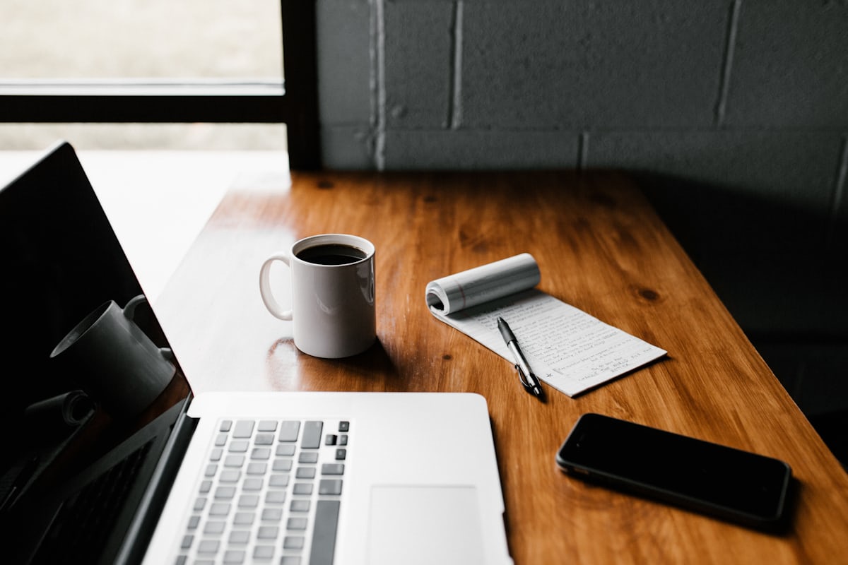 A wooden desk holding an open laptop, a notebook, a pen and a cup of coffee.